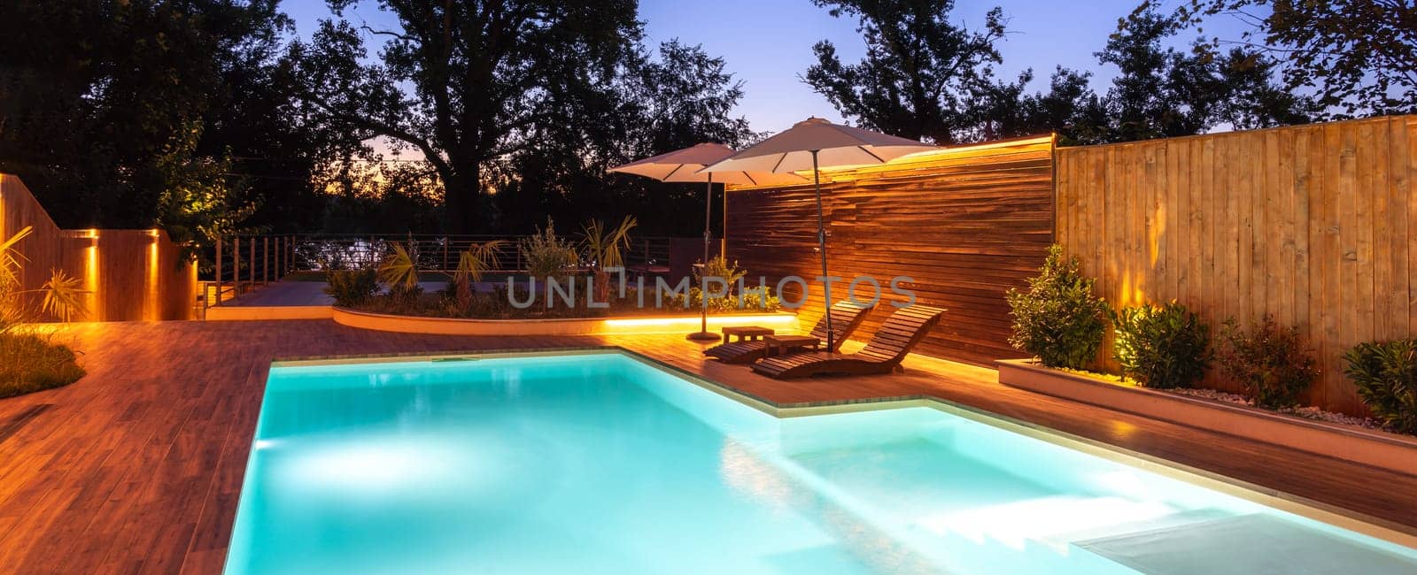A nighttime scene of a pool and deck illuminated with warm lighting, surrounded by lush greenery, creating a luxurious and tranquil setting. Concept of relaxing outdoor atmosphere and elegant evening leisure