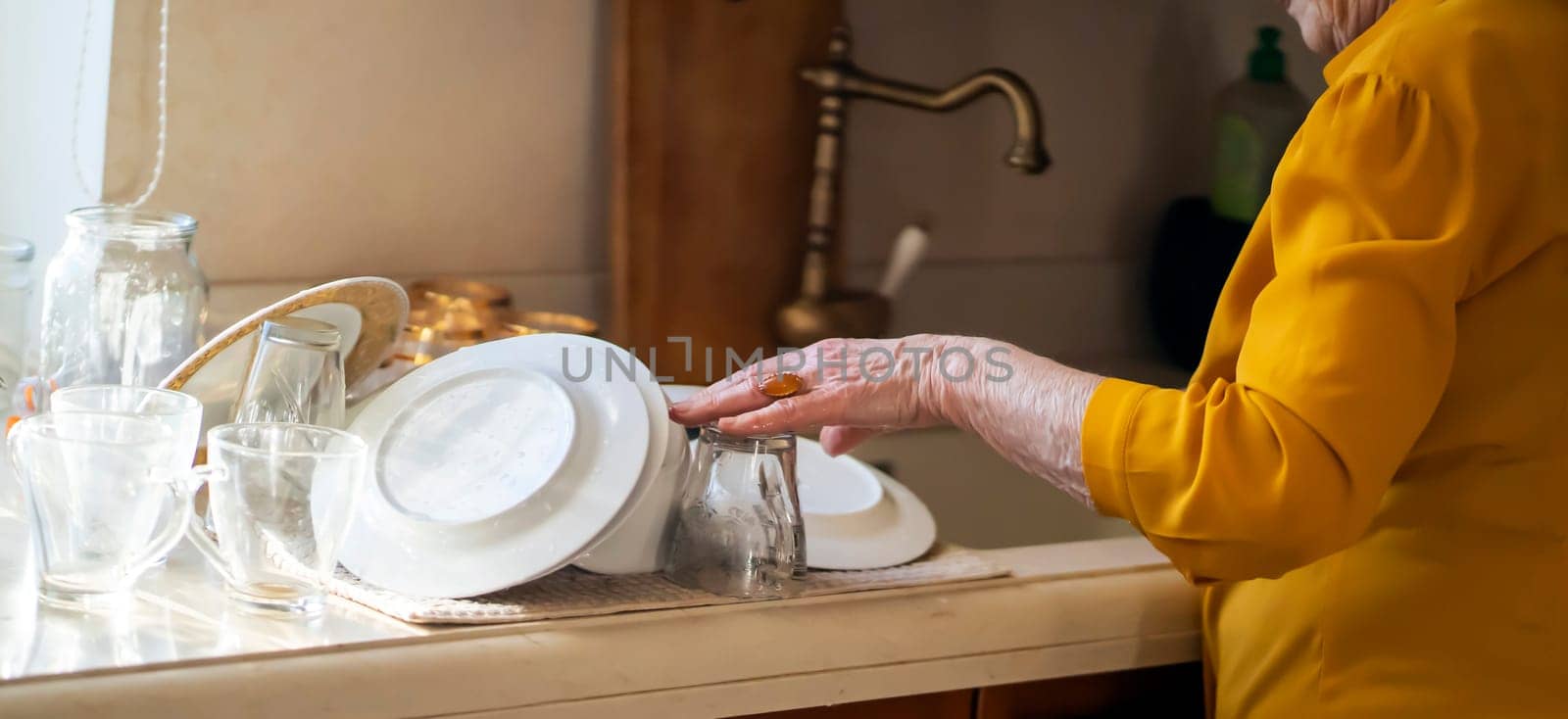 Elderly retired gray-haired woman having a good time in her house, active in her old 80+ age, washing dishes in a cozy kitchen, cleaning. A happy grandmother in a yellow blouse, young at heart.