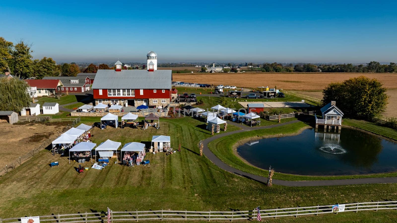 Ronks, Pennsylvania, USA, October 20 2024 - Visitors enjoy a lively autumn market set in front of a large red barn, featuring white tents and a serene pond. The clear blue sky enhances the cheerful ambiance of the day.
