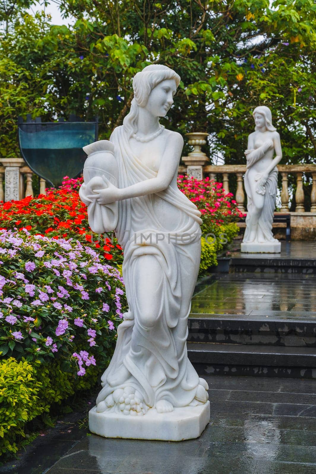Sculptures of mythological goddesses with a vase in their hands made of white stone in a blooming garden