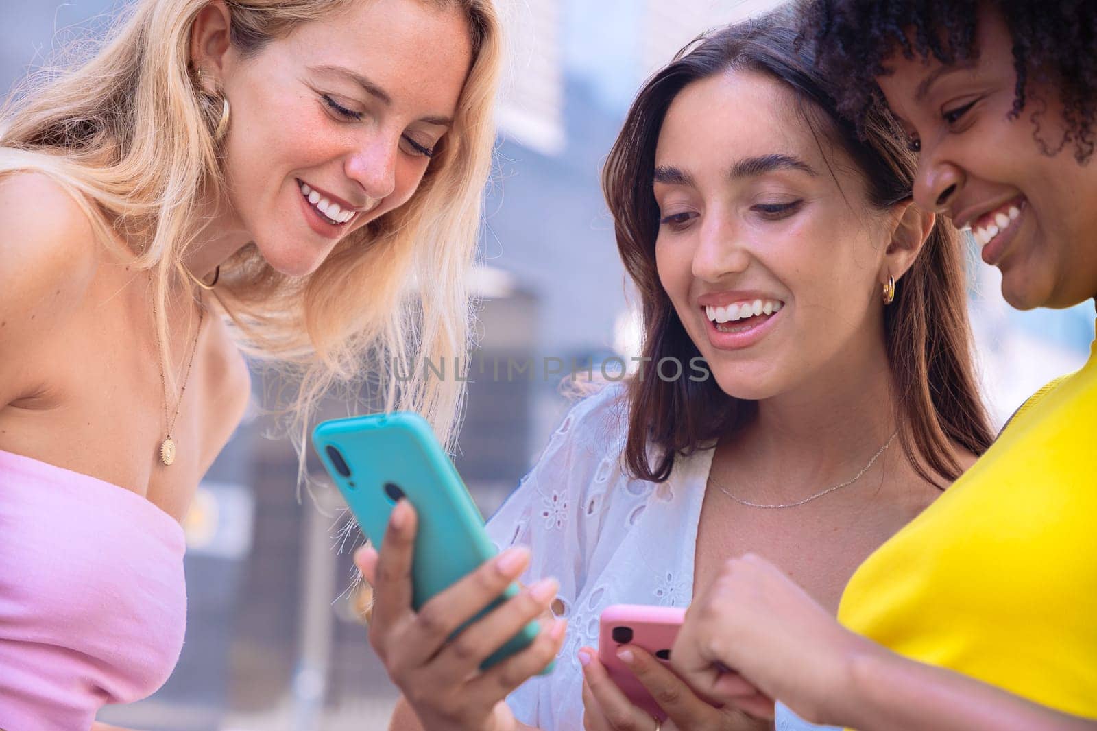 Three cheerful young women gathers around a red smartphone, reacting with excitement. They are dressed casually, standing outdoors in a modern urban environment, enjoying a fun moment together.