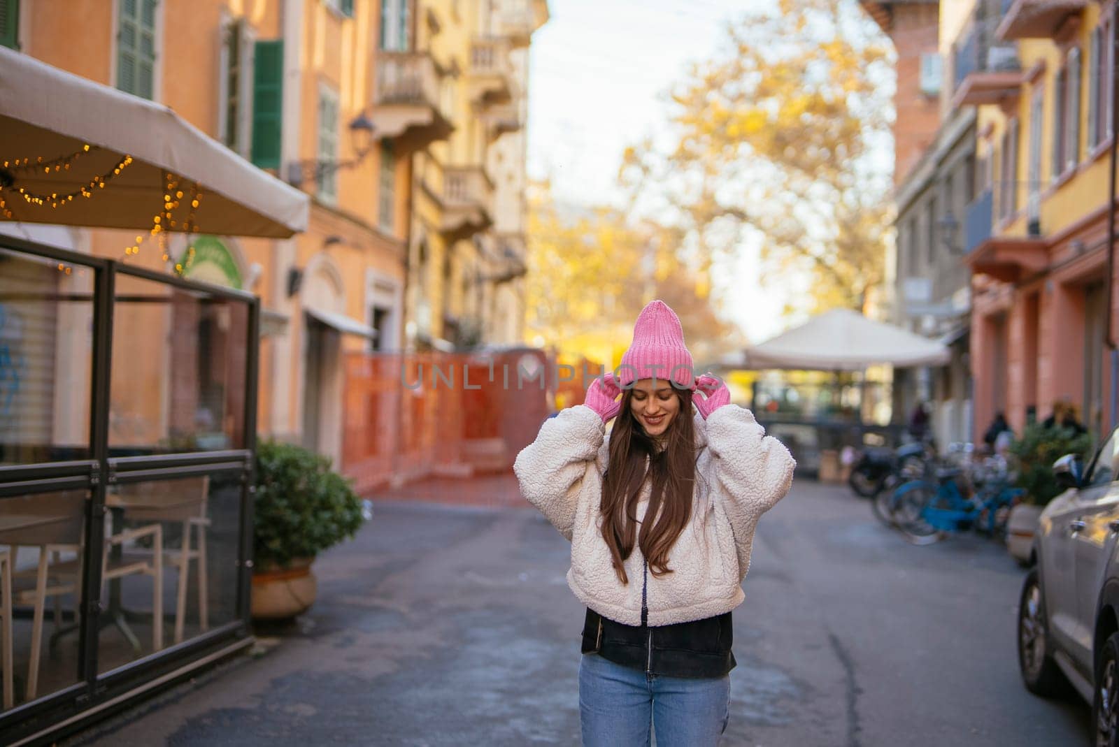 A young woman wearing a vibrant pink hat poses happily on a charming city street filled with life and color