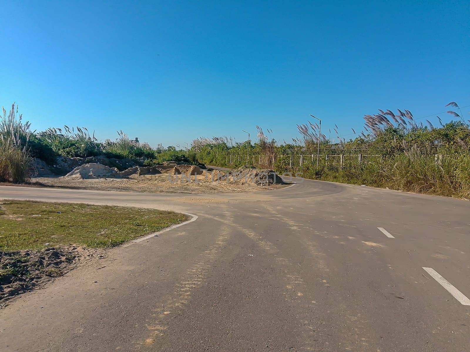 A remote, sunlit road curves through a rural landscape, flanked by tall grasses rustling in the breeze. Mounds of excavated earth and patches of dirt hint at recent construction activity. The clear blue sky evokes tranquility, contrasting with the signs of human disruption.