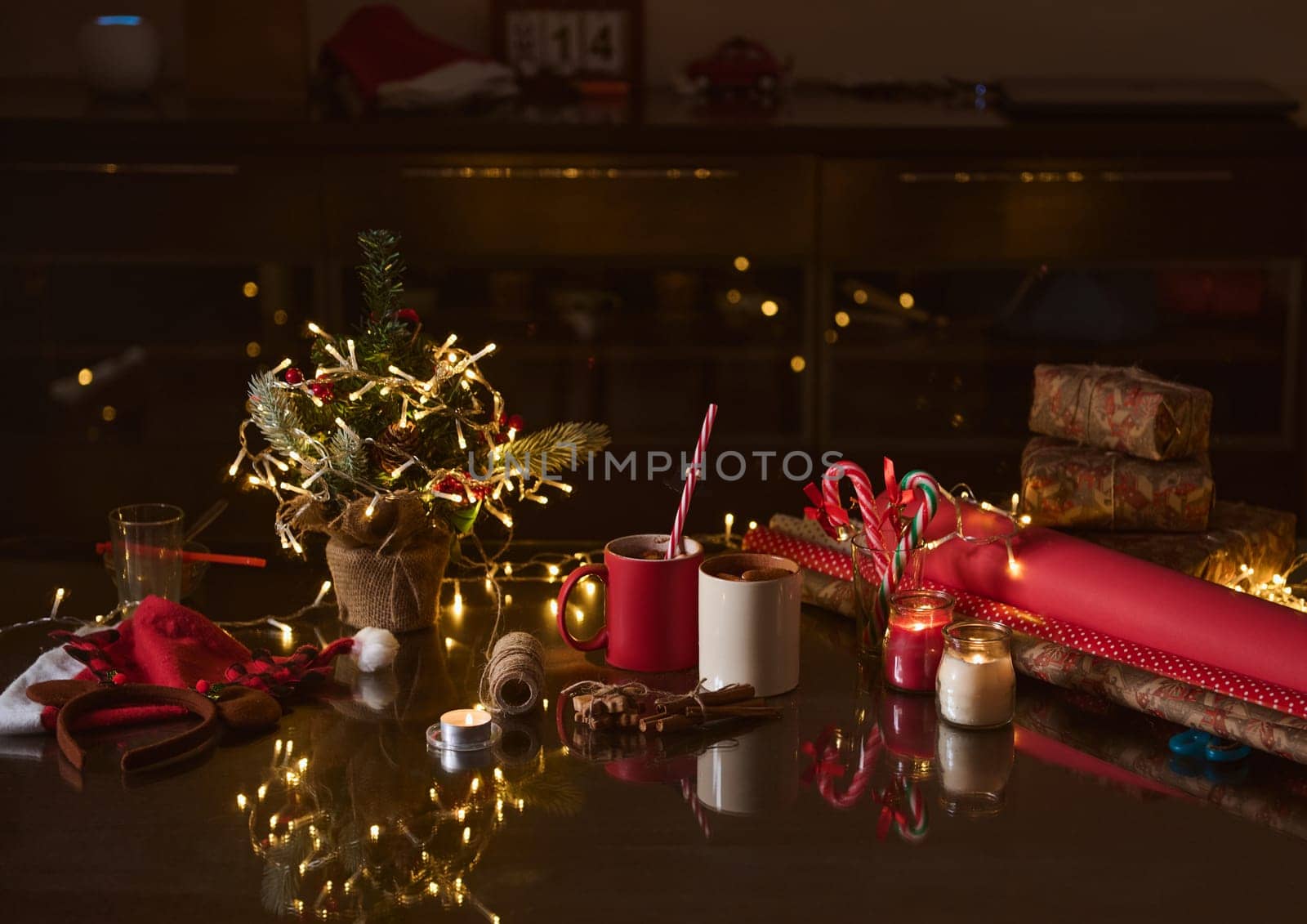 Festive table setting featuring Christmas decor, mugs of hot chocolate, and gift wrapping supplies in a warm, inviting atmosphere enhanced by fairy lights.