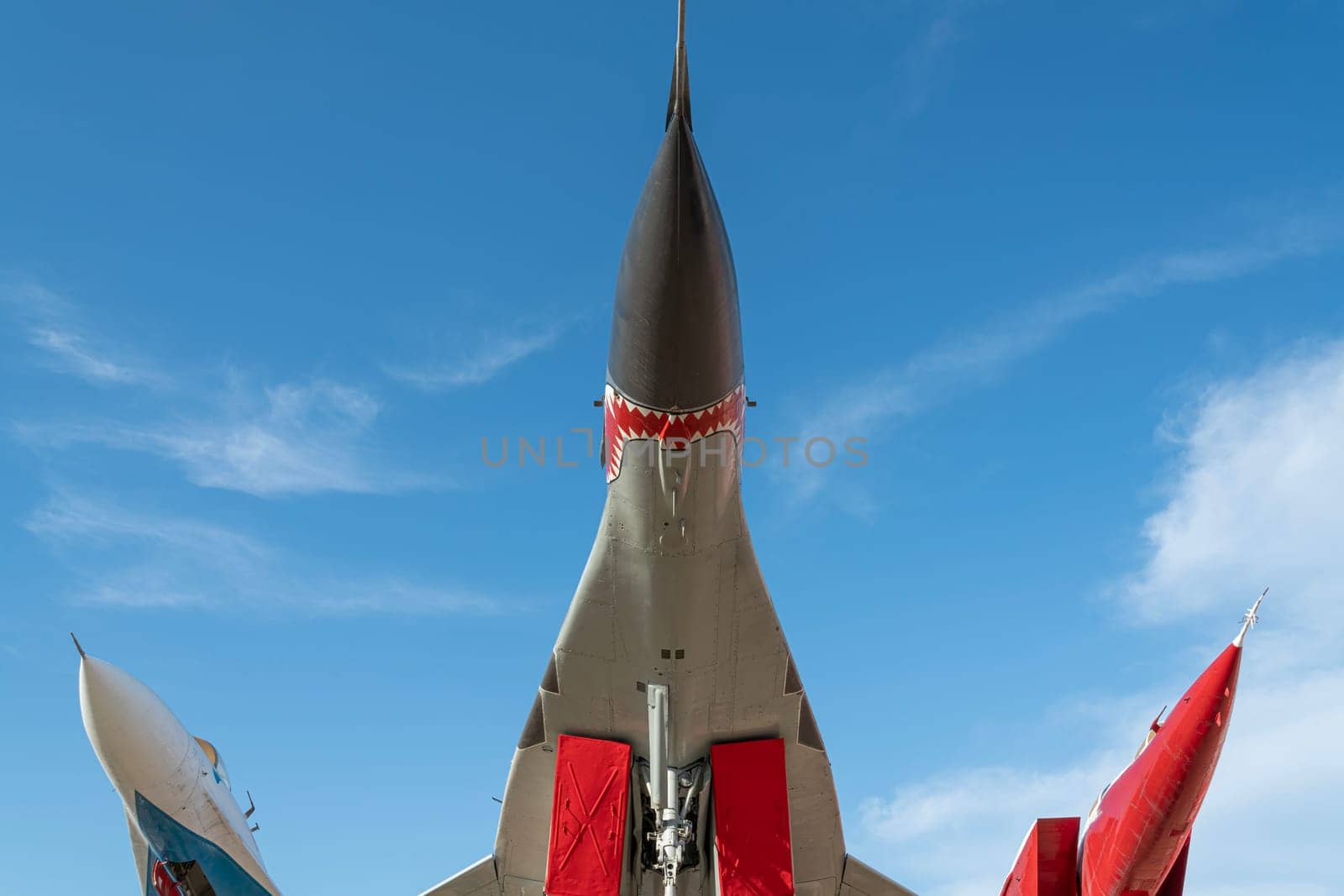 A fighter jet is showcased outdoors, with vibrant red detailing and a striking design. Bright blue skies provide a perfect backdrop for this military aircraft exhibit during the day.