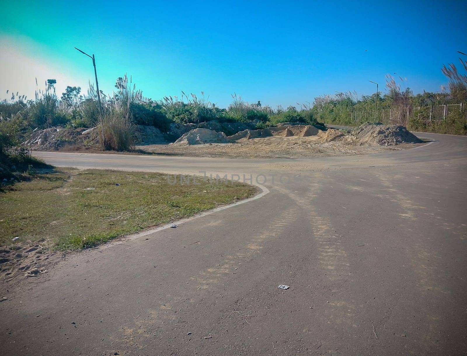 A desolate road curves gently beside a large, barren mound of earth, hinting at recent construction or excavation. Under a clear blue sky, the scene is stark and abandoned, with tall, dry grasses and scattered lamp posts lining the roadside. The mood is one of quiet, forgotten stillness.