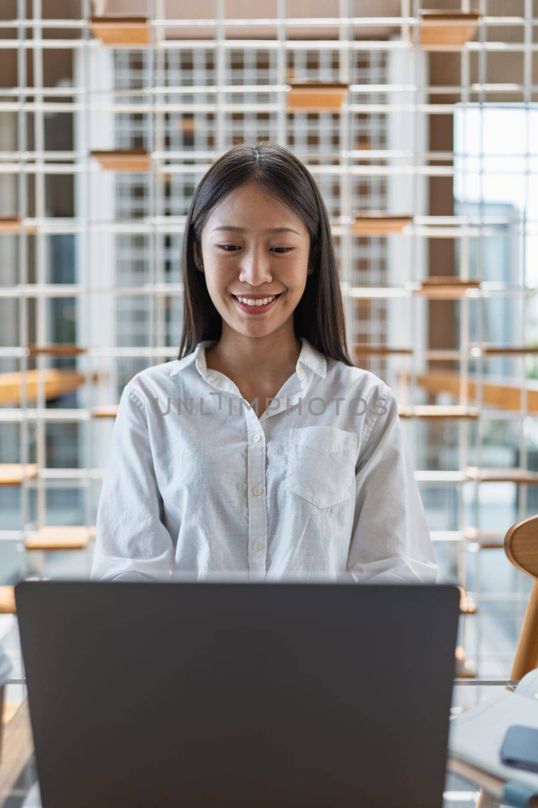 A young woman sits at a modern desk, smiling while using her laptop, illustrating a scene of productivity, positivity, and contemporary workspace design in a bright environment.