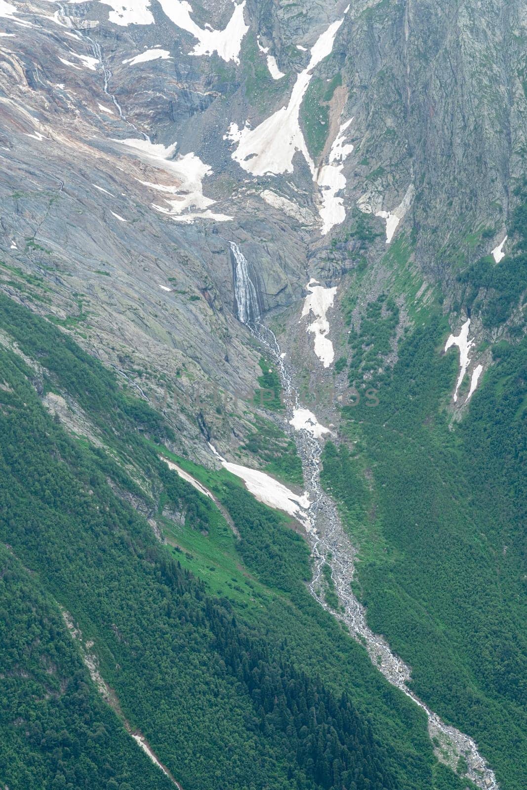 A waterfall flows down steep rocks in a mountainous area. Lush green vegetation surrounds the rocky terrain, creating a serene and tranquil atmosphere in this natural setting.