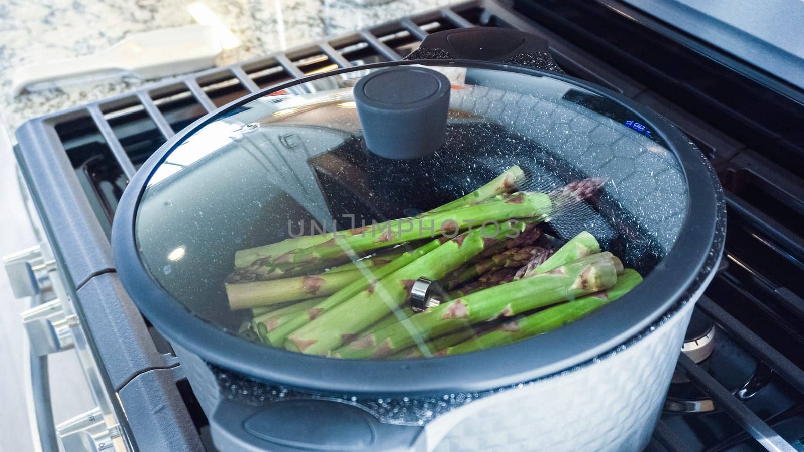 In a modern white kitchen, large organic asparagus is being steamed in a sizable cooking pot.
