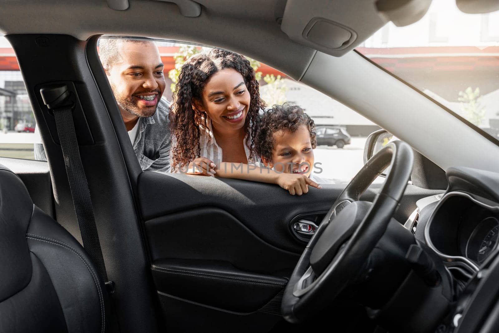 Friendly african american family choosing new car at dealership center parking spot. Excited black mom, dad and preteen child son looking inside new automobile and smiling. Auto buying, transportation