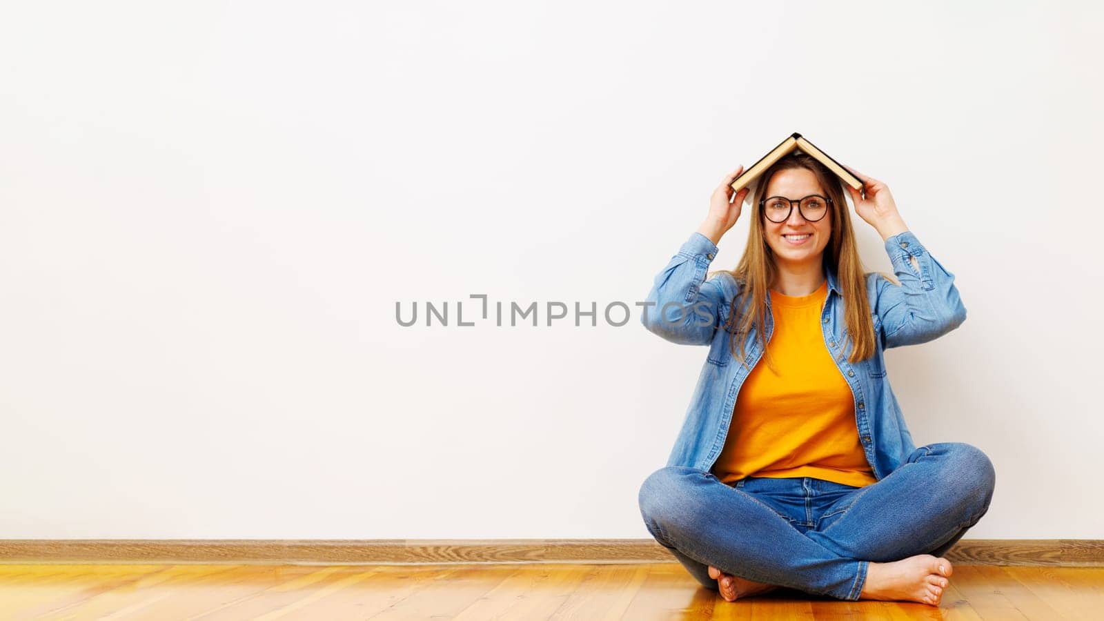 Woman in glasses holding opened book on head while sitting on the floor near white wall with copy space. Concept of education, organization of courses, conducting training classes