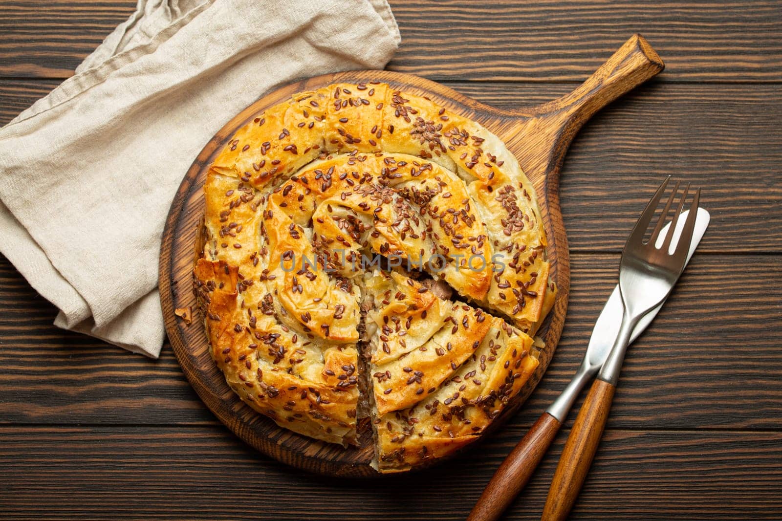 Burek with cut piece made of filo dough with filling on cutting board, dark brown wooden rustic background top view. Traditional savoury spiral pie of Balkans, Middle East and Central Asia.