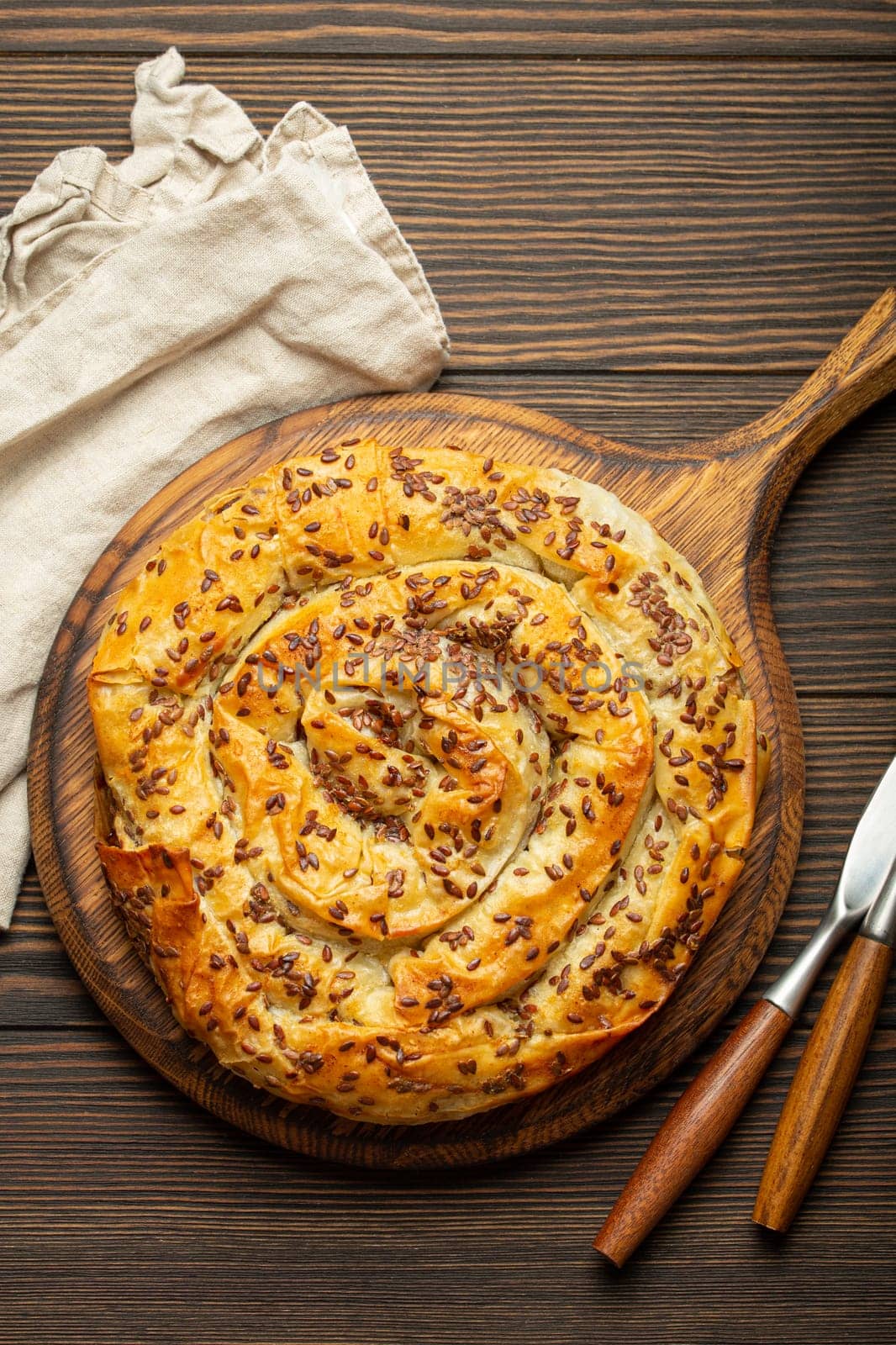 Burek made of phyllo dough with filling on cutting board, dark brown wooden rustic background top view. Traditional savoury spiral pie of Balkans, Middle East and Central Asia.