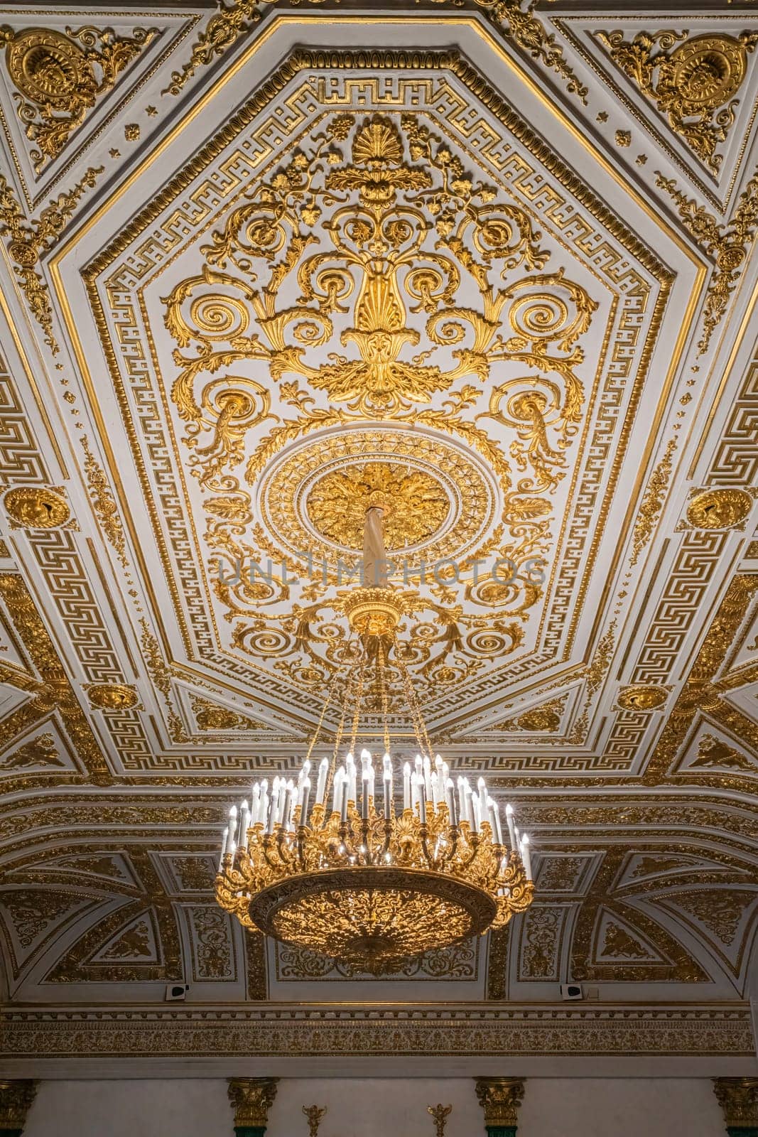An ornate hall features a stunning chandelier beneath a beautifully detailed ceiling, showcasing elaborate gold leaf patterns and classical architectural design elements.
