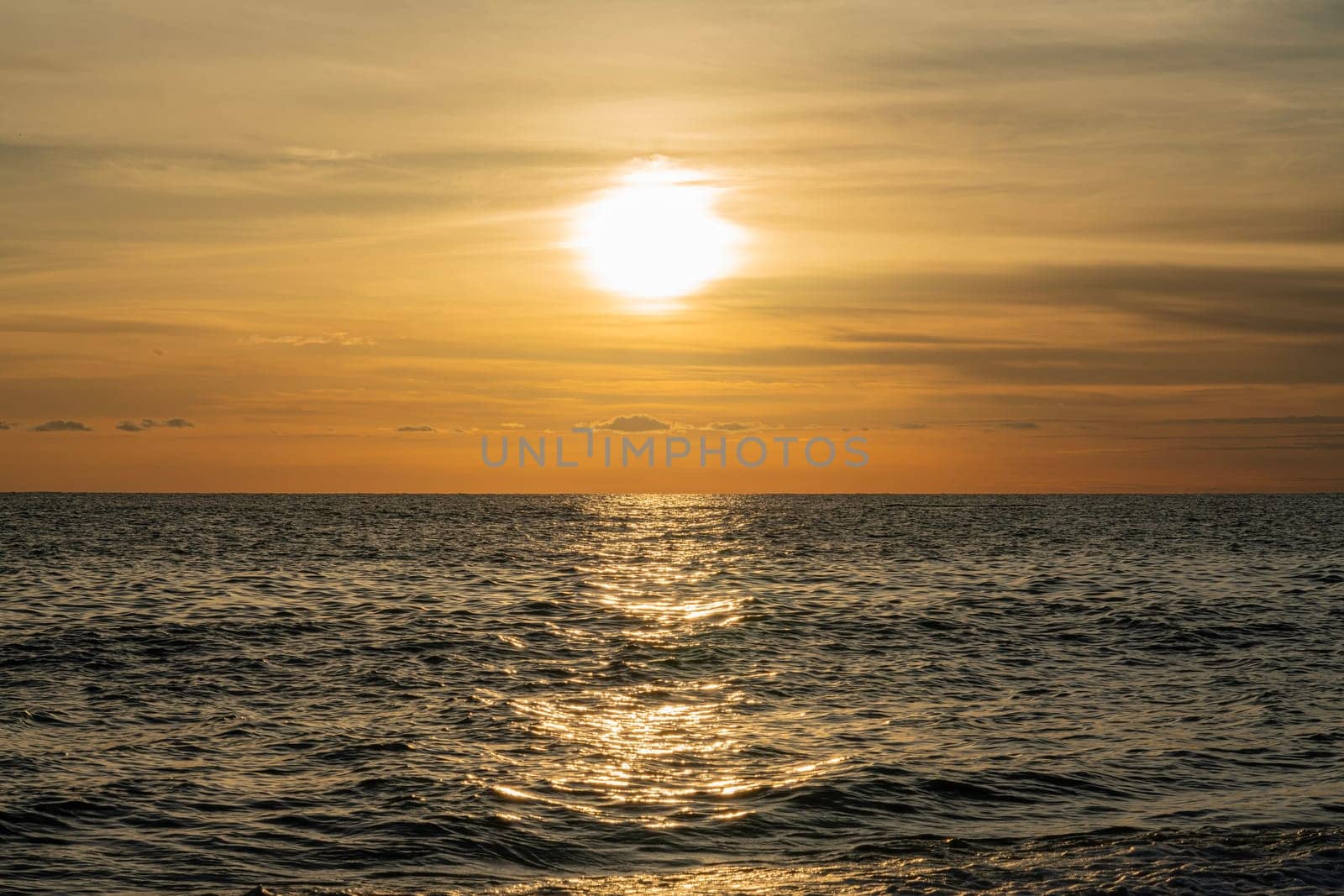 The sun sets on the horizon, casting a golden glow across the ocean surface. Gentle waves ripple beneath the warm light, creating a tranquil and peaceful atmosphere by the water.