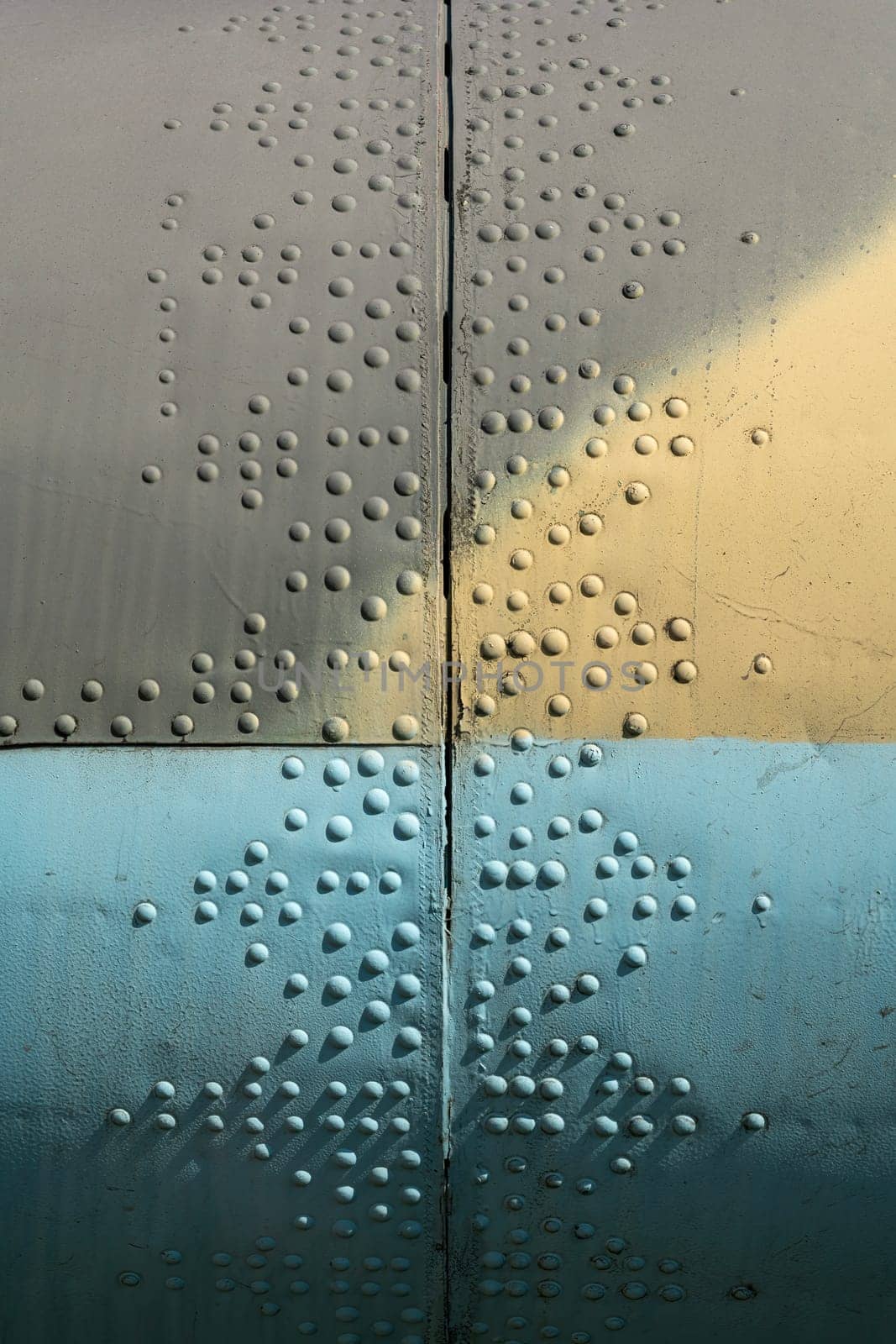 Close-up view of a textured industrial surface featuring colorful sections and rivets. The image highlights the interplay of light, shadow, and color on the metal.