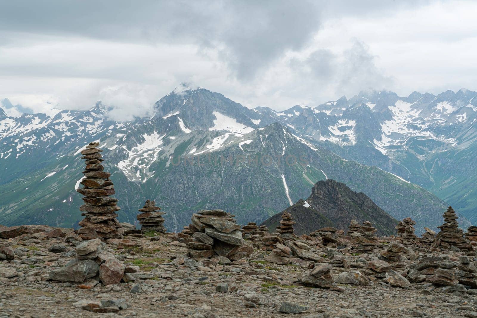 Cairns of stacked stones are arranged on a rocky mountain top, surrounded by majestic peaks and low-hanging clouds. The green valleys contrast beautifully with the snowy mountaintops.