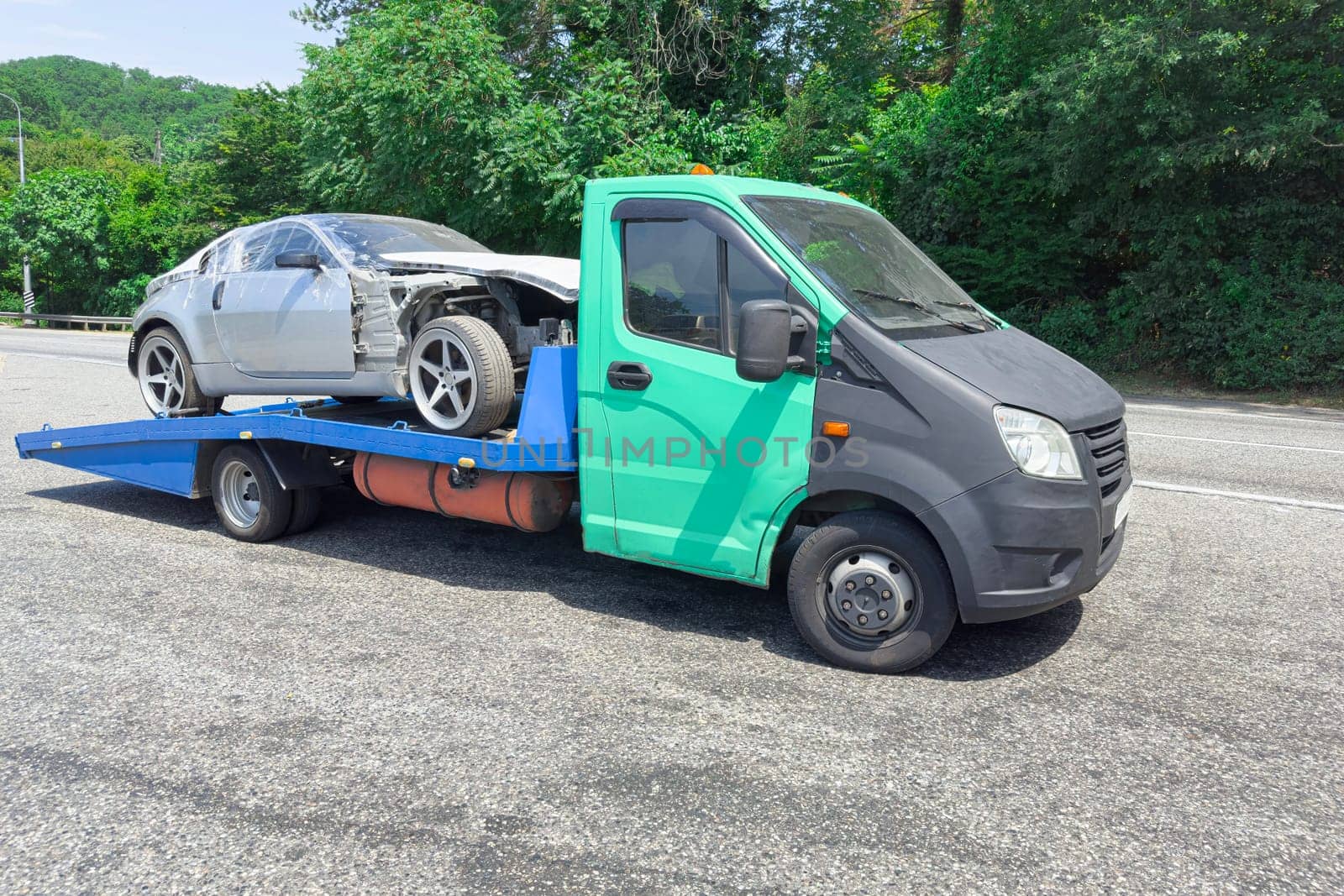 A tow truck carries a silver car with damage on a blue flatbed along a rural road. Lush greenery surrounds the area, indicating a sunny day.