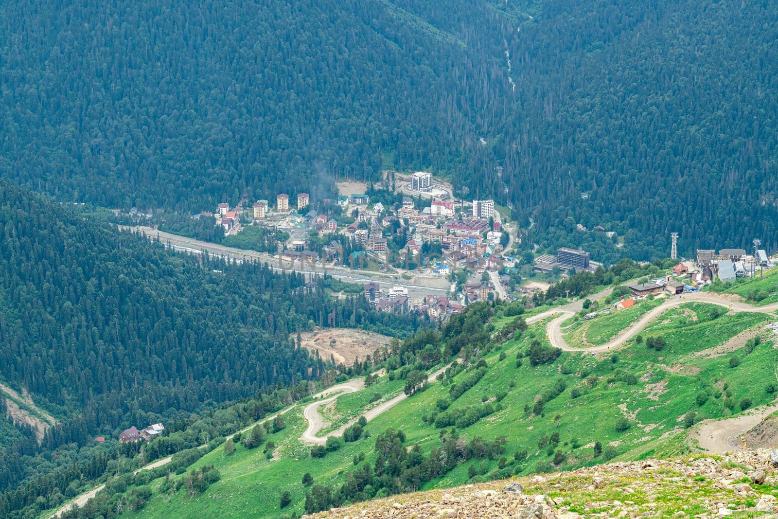 A picturesque view of a charming mountain village surrounded by dense forests and rolling hills. Winding roads traverse the vibrant green landscape under a clear summer sky.