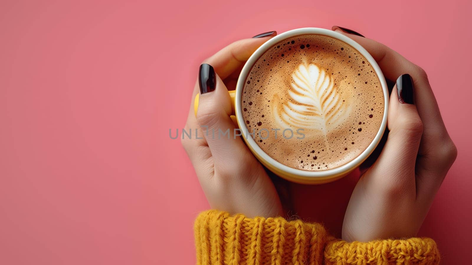 Close-up of hands in yellow knit sweater holding a cappuccino cup with beautiful latte art on a soft pink background.