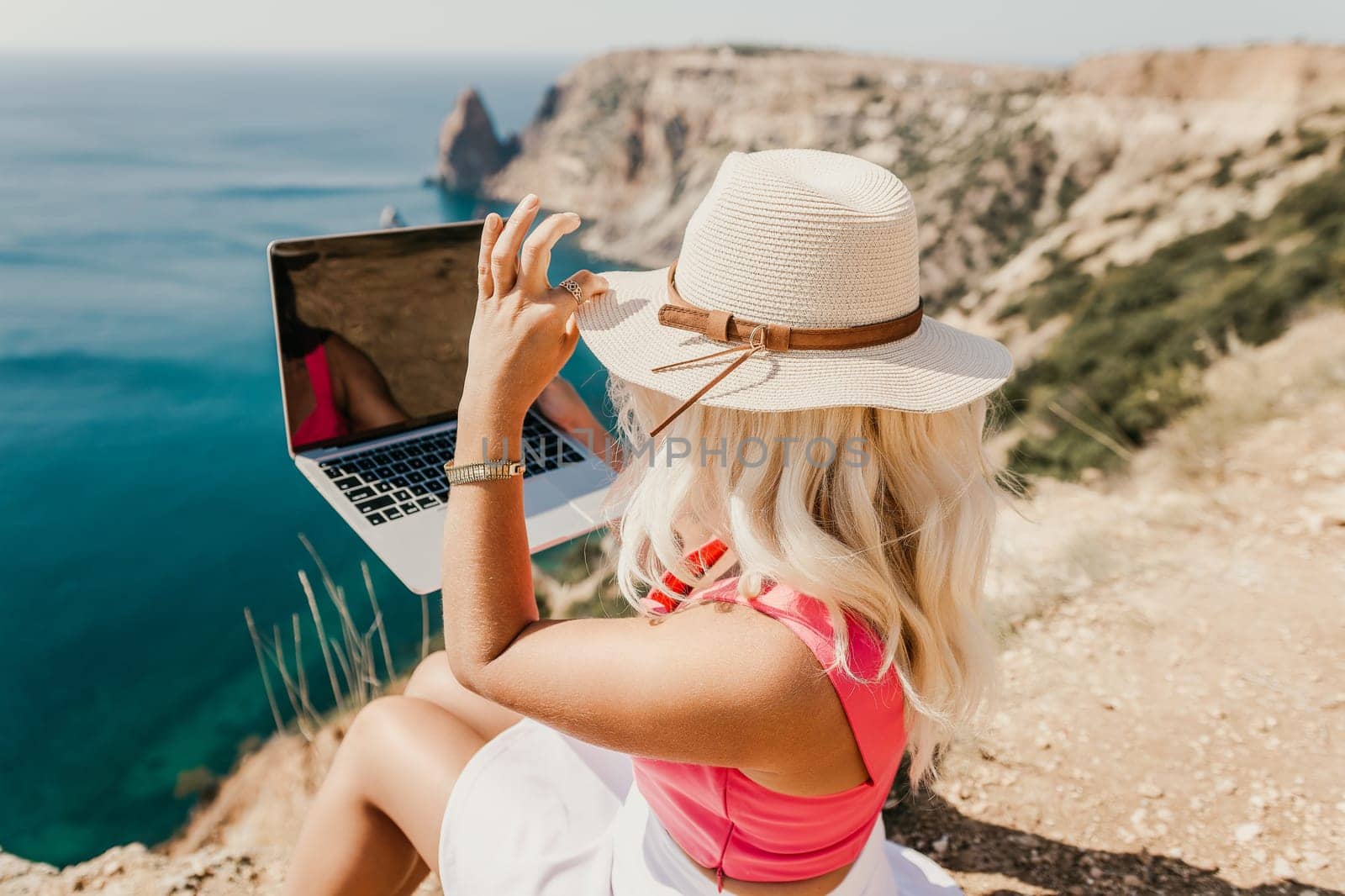 A woman is sitting on a rocky beach with a laptop in front of her. She is wearing a pink top and a straw hat. Concept of relaxation and leisure