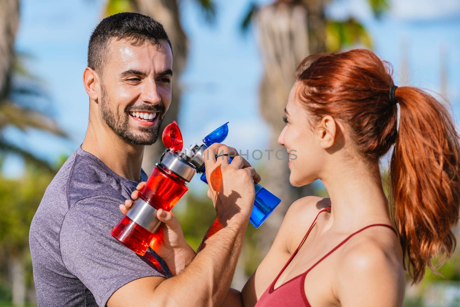 man and woman smile and interact while holding colorful water bottles outdoors. They are dressed in sporty attire, enjoying a sunny day and staying hydrated during their fitness activity in a park setting.