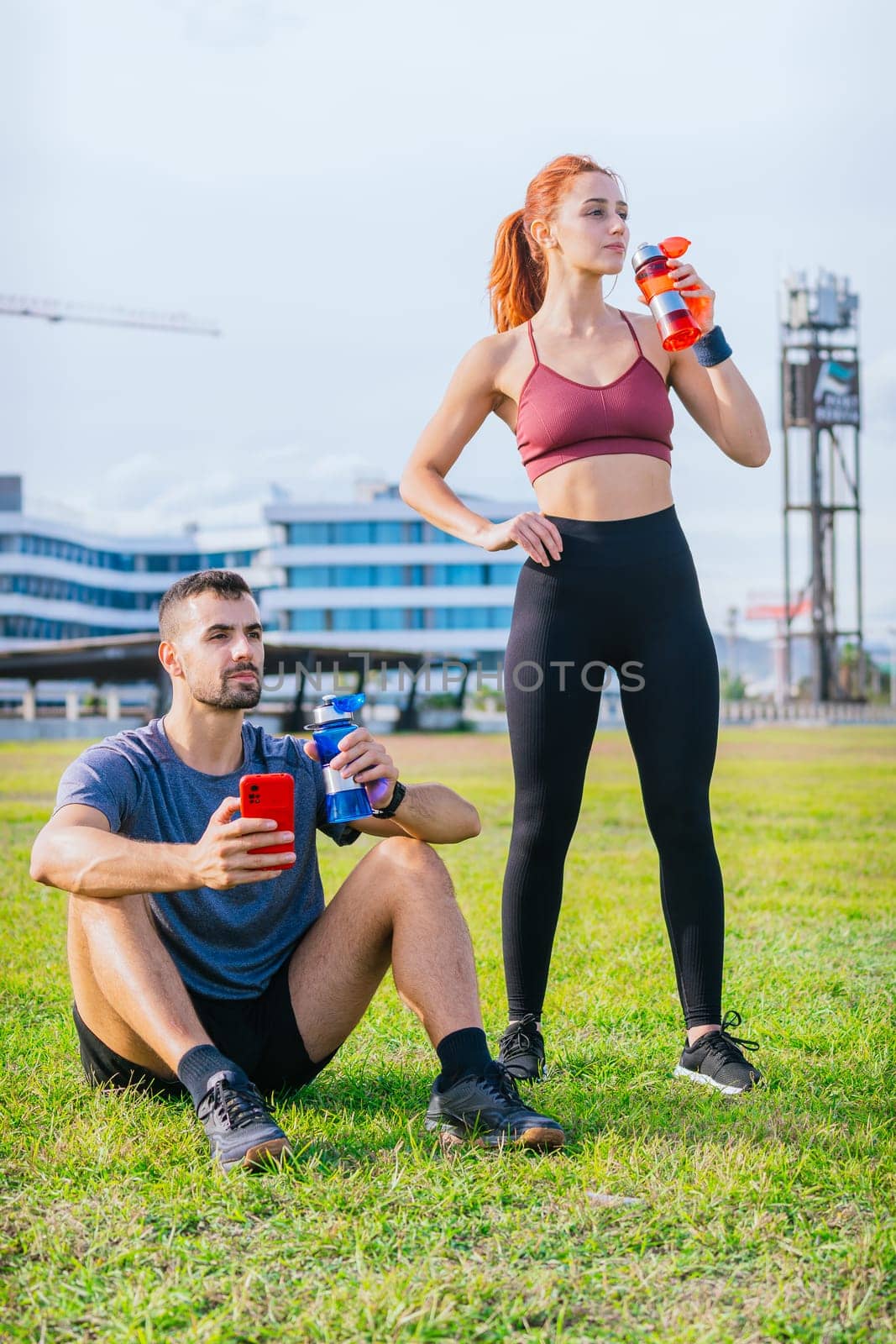 man sits on the grass holding a red smartphone and blue water bottle, while a woman stands nearby drinking from a red bottle. Both are dressed in sporty attire, relaxing in a sunny urban park after exercising.