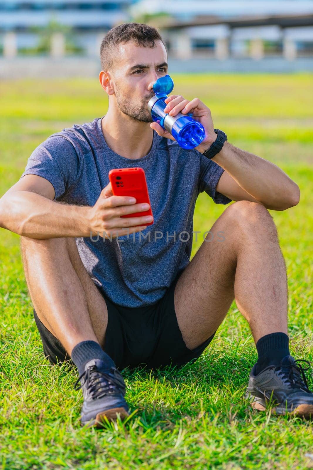 A fit man sits on the grass in a park, drinking water from a blue bottle while holding a red smartphone. He wears sporty attire, resting after exercise on a sunny day in a natural outdoor setting.