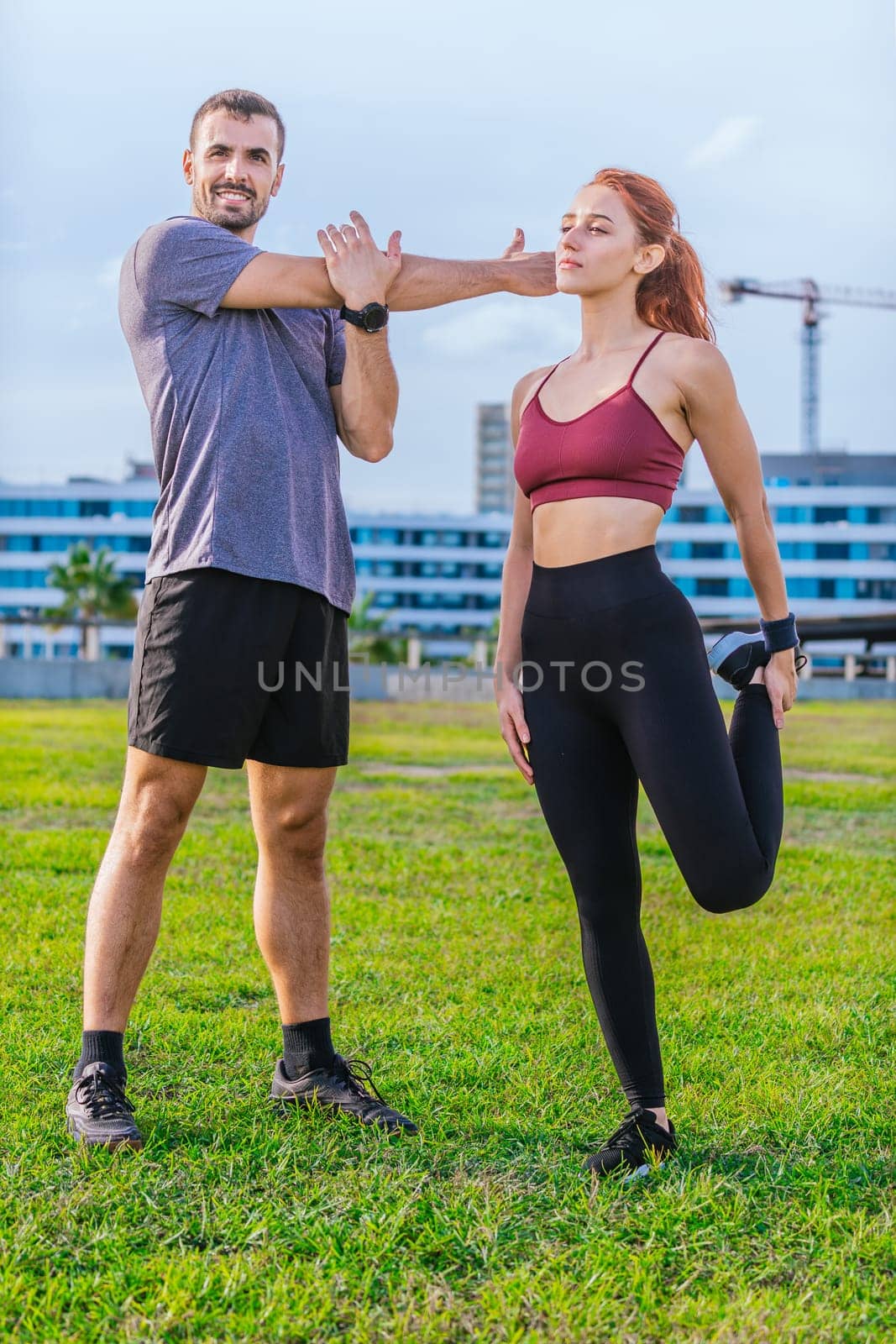 man and woman stretch their muscles during a workout session in an urban park. The man raises his arms while the woman lifts her knee, both dressed in sporty attire and enjoying a sunny, active day.