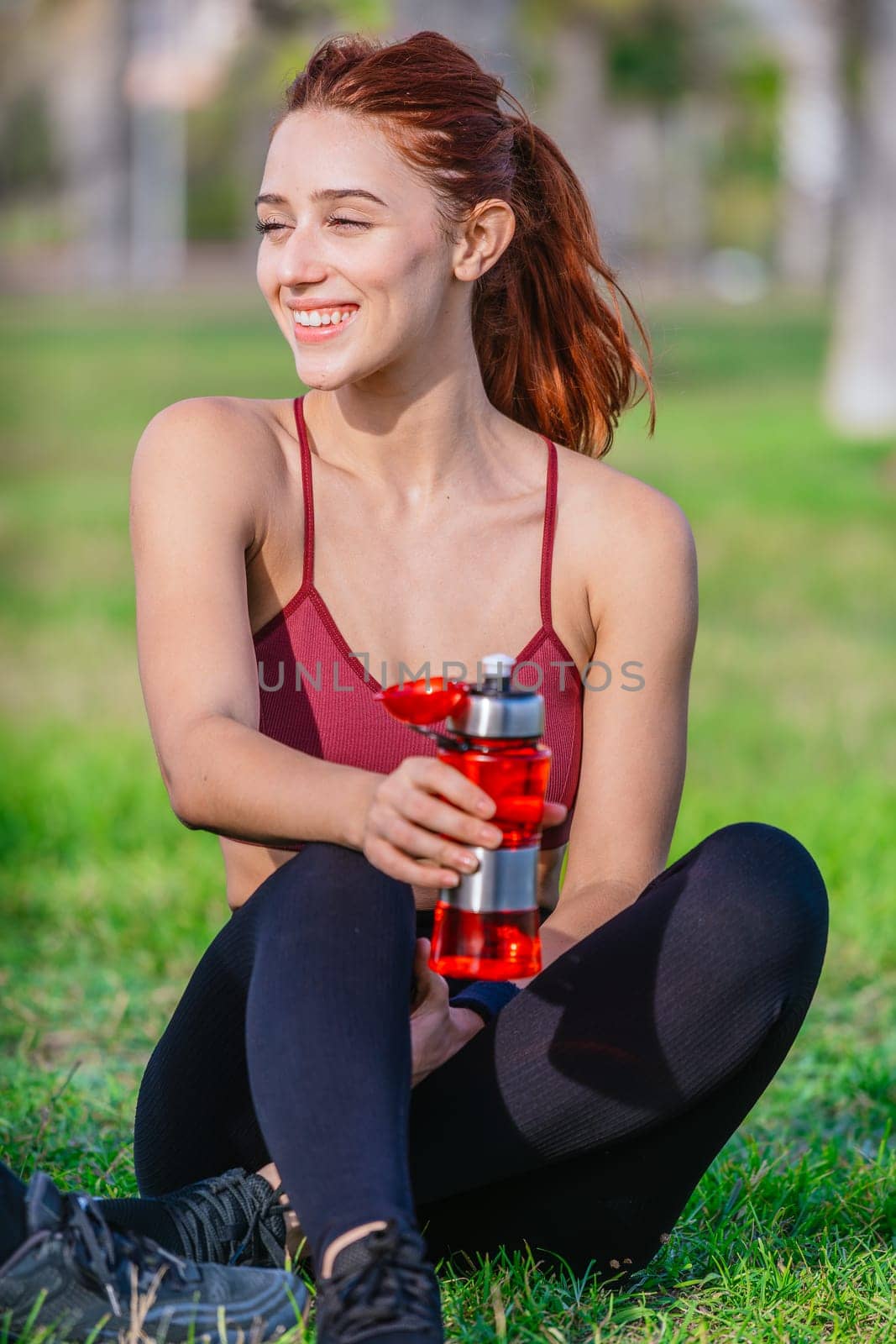 A fit young woman with red hair sits on the grass holding a red water bottle. She is dressed in sporty attire, smiling and enjoying a sunny day outdoors in a natural park setting after exercising.