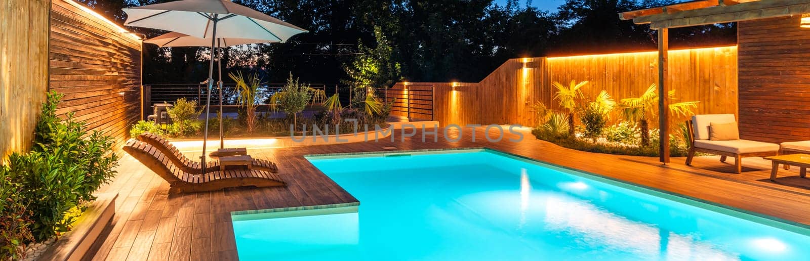 Luxury swimming pool with wooden deck surrounded by lush plants and warm ambient lighting at night. Concept of a modern outdoor relaxation area for leisure and recreation