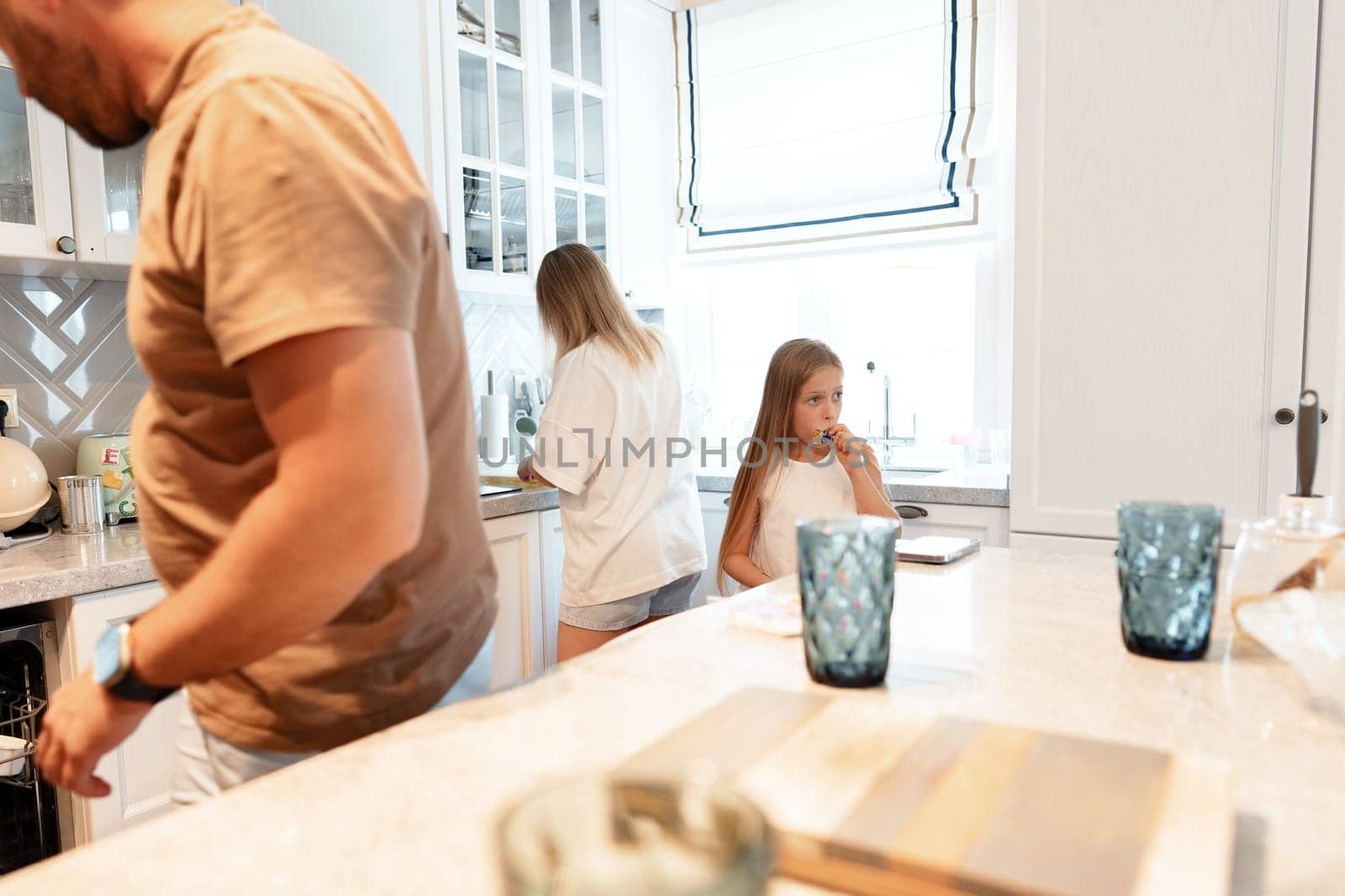A family is in their kitchen. The father is loading the dishwasher, while the mother is washing dishes. The daughter is sitting at the counter eating a snack.