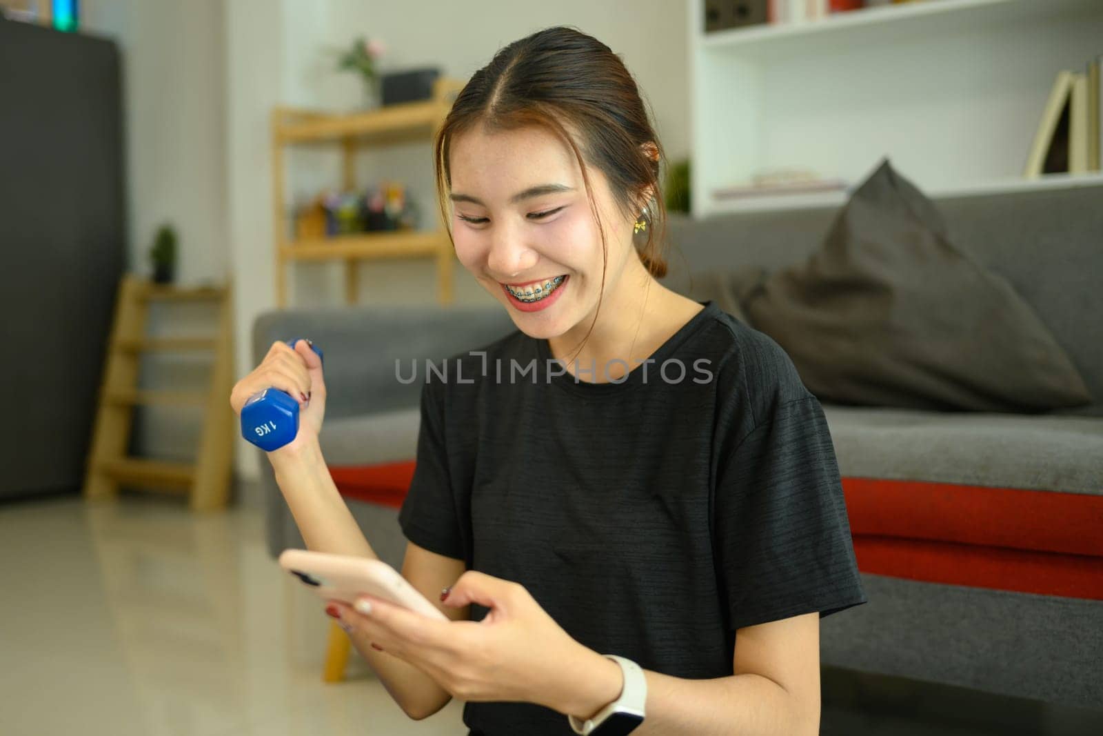 Smiling young woman lifting a light dumbbell and using smartphone in living room.