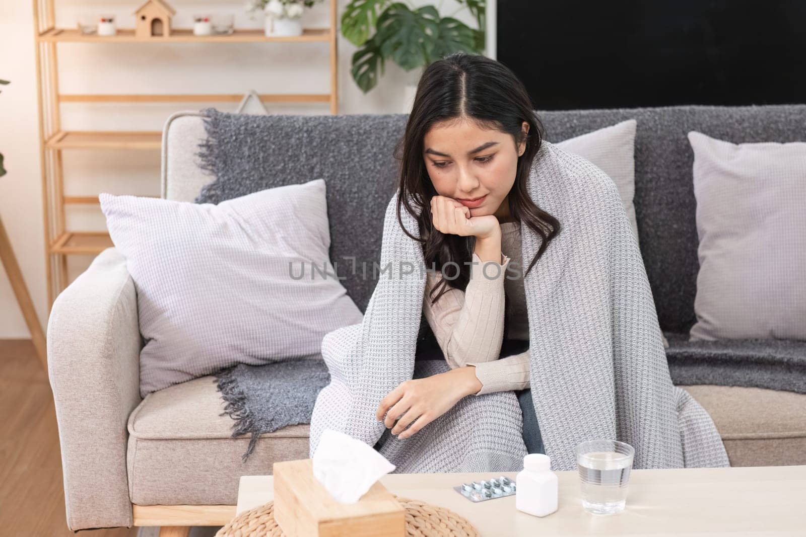 A woman feeling sick and unwell wrapped in a blanket contemplating her wellness in her cozy living room.
