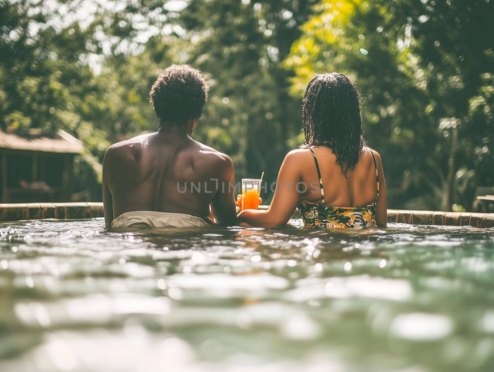 Back view of a couple sitting in a pool, holding drinks, and enjoying a relaxing moment surrounded by lush tropical greenery.