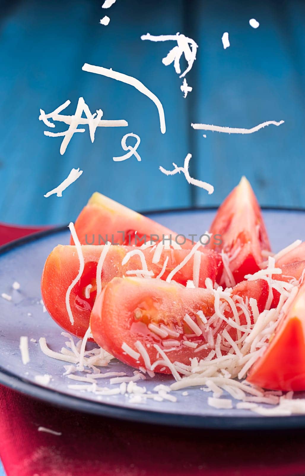 Shredded white cheese being sprinkled onto tomato wedges on a plate.