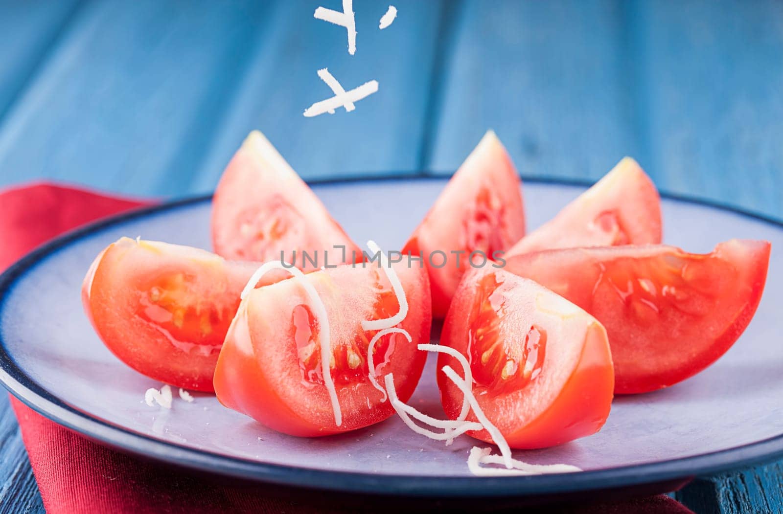 Shredded white cheese being sprinkled onto tomato wedges on a plate.