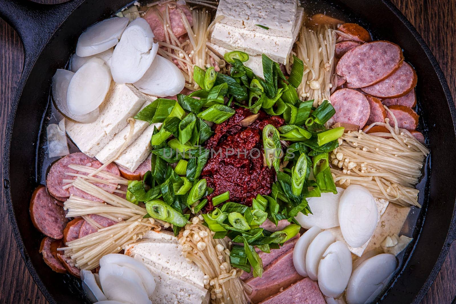 A close up of a modern Korean dish using sausage, tofu, onions, mushrooms, and red pepper paste.