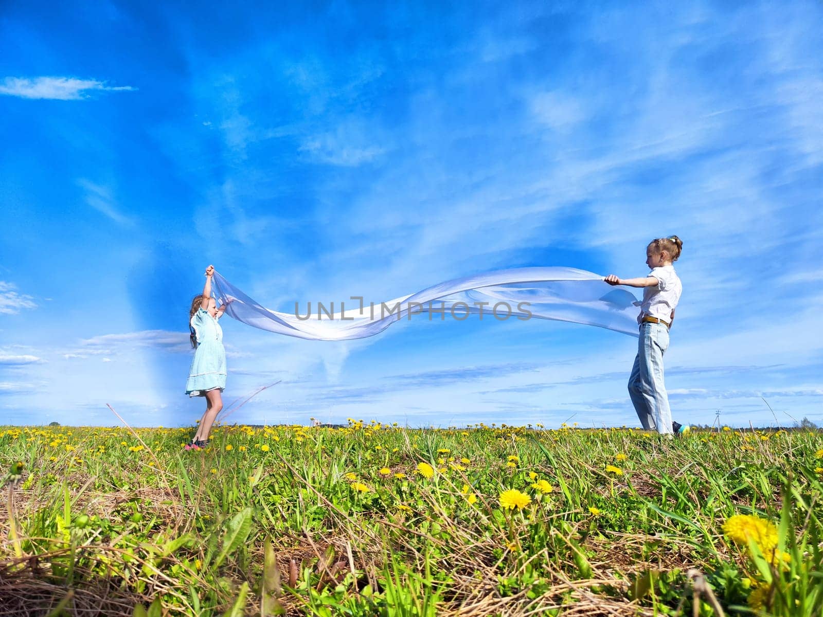 A child and an adult happily wave colorful fabric in a vibrant green field filled with wildflowers under a clear blue sky.