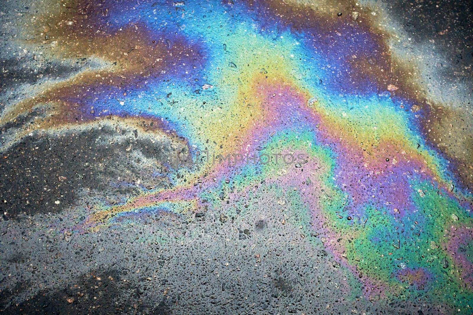 A colorful chemical sheen appears on the asphalt road from a gasoline or oil spill.