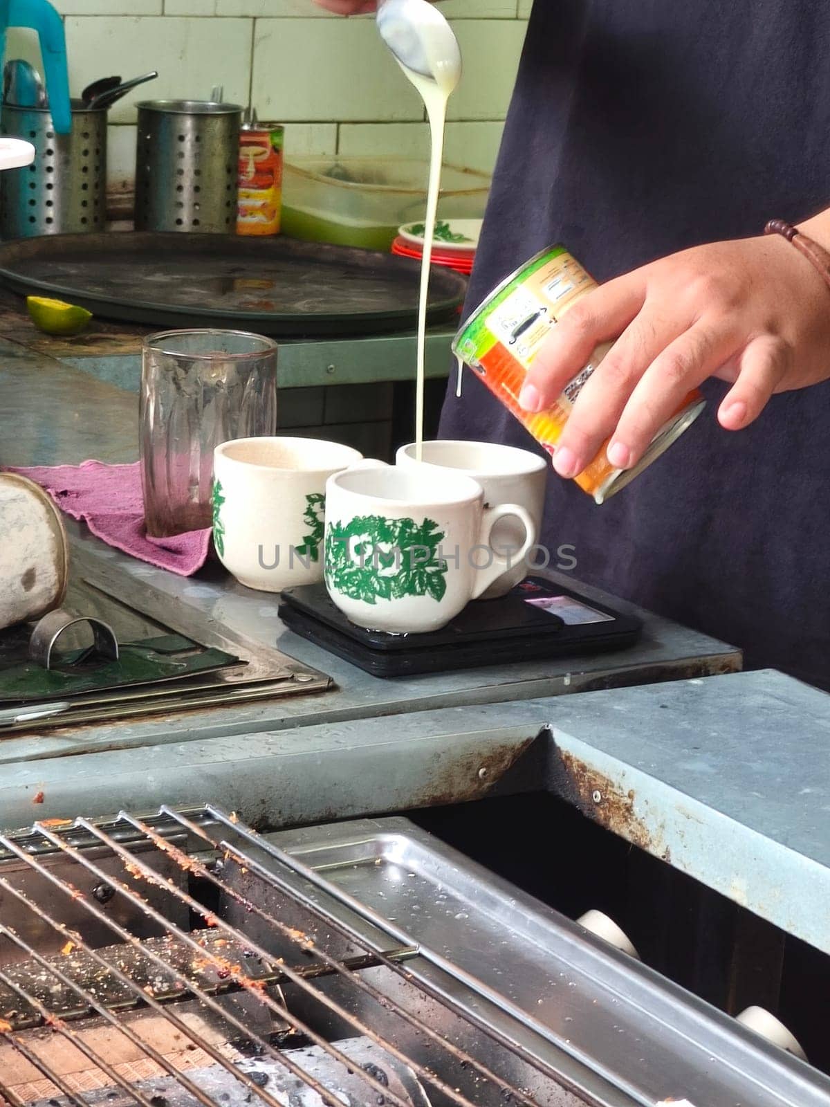 A barista pours condensed milk from a can into a small cup on a scale, likely preparing a coffee drink.