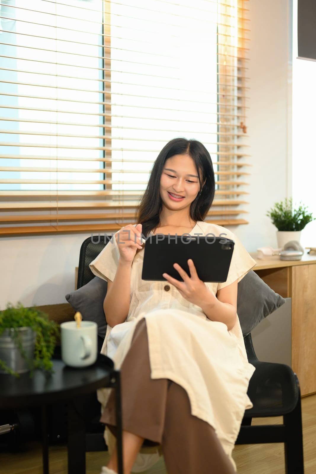 Young woman in casual clothing using digital tablet on chair in living room.
