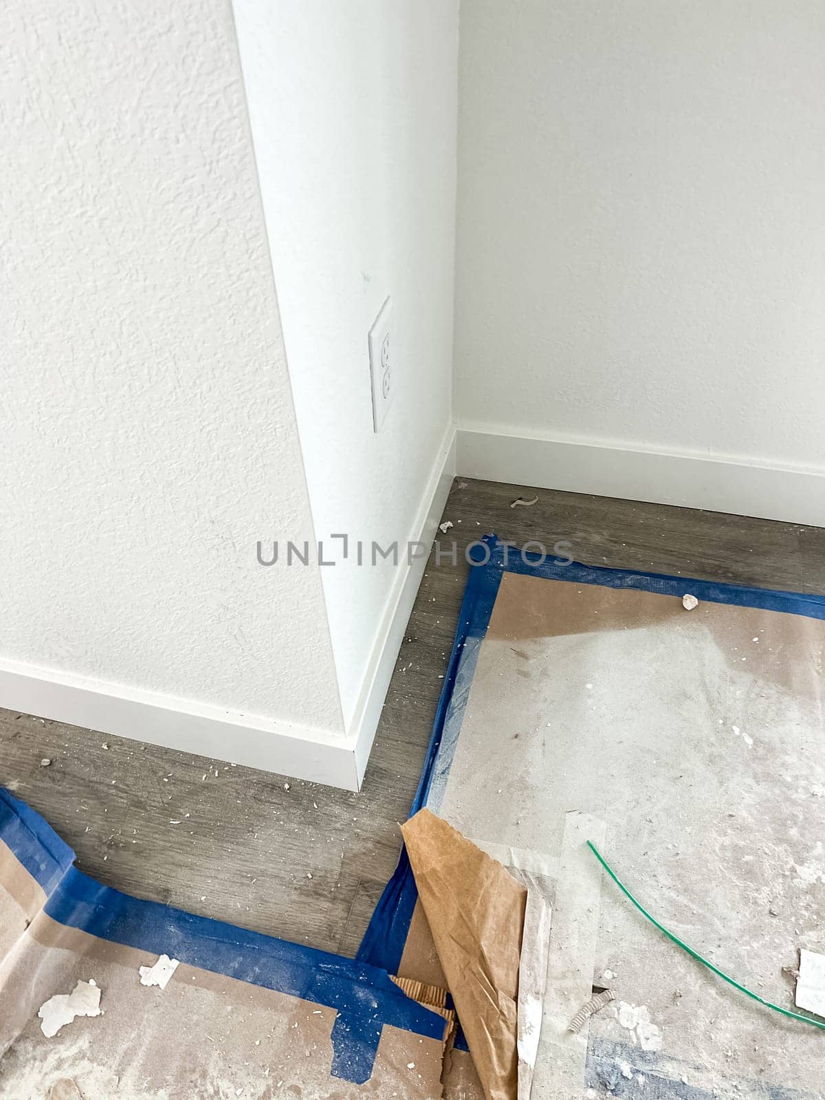 Parker, Colorado, USA-January 11, 2023-A close-up view of a corner in a new construction home featuring newly installed baseboards, a finished white wall, and flooring partially covered with protective paper and painter tape. Construction debris and materials are visible, indicating ongoing work.