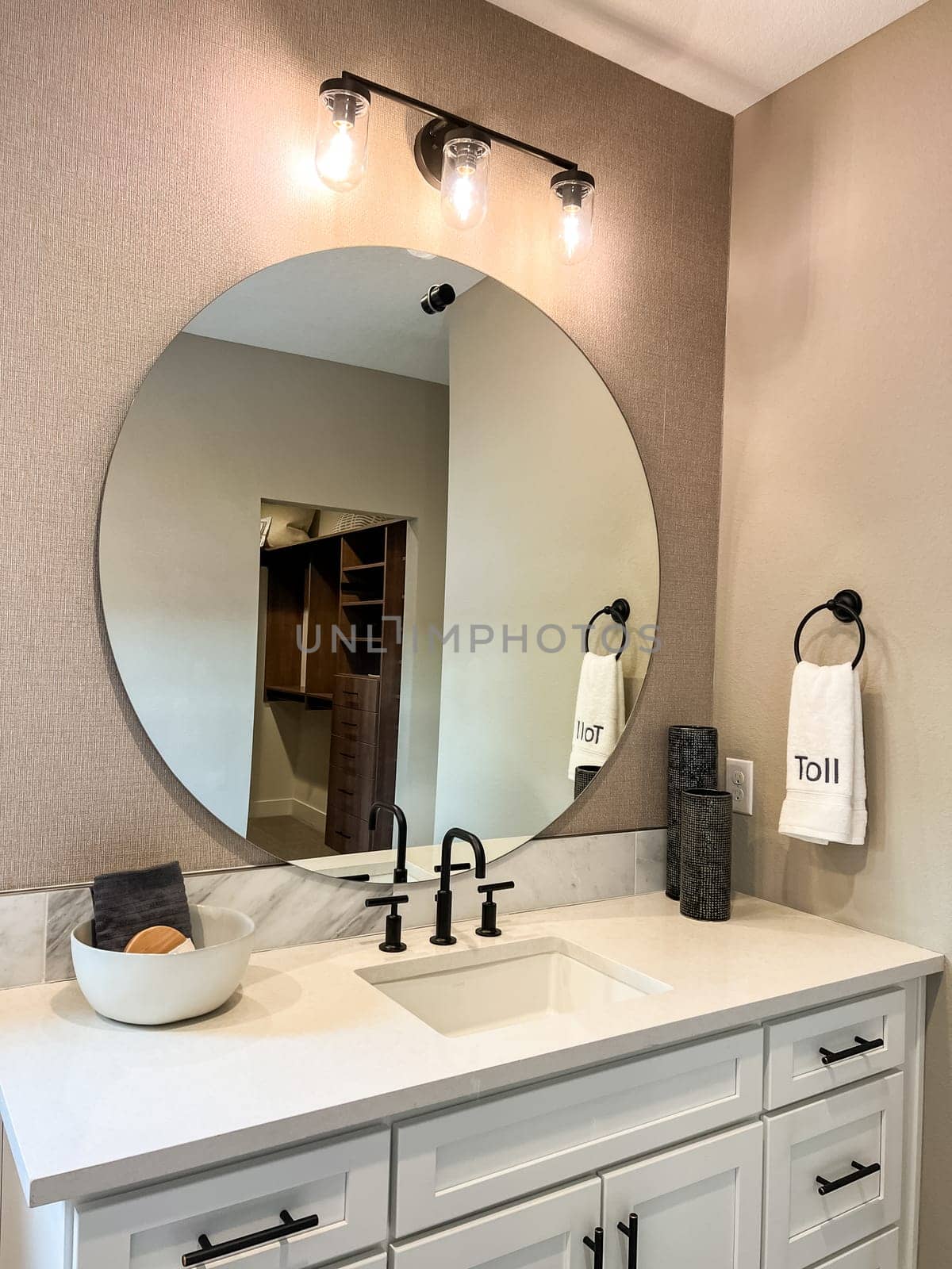 Parker, Colorado, USA-January 11, 2023-Stylish bathroom vanity featuring a round mirror, black fixtures, and a bright light fixture above. The vanity has a white countertop with a sleek undermount sink and black faucet. A reflection shows part of a walk-in closet in the background.