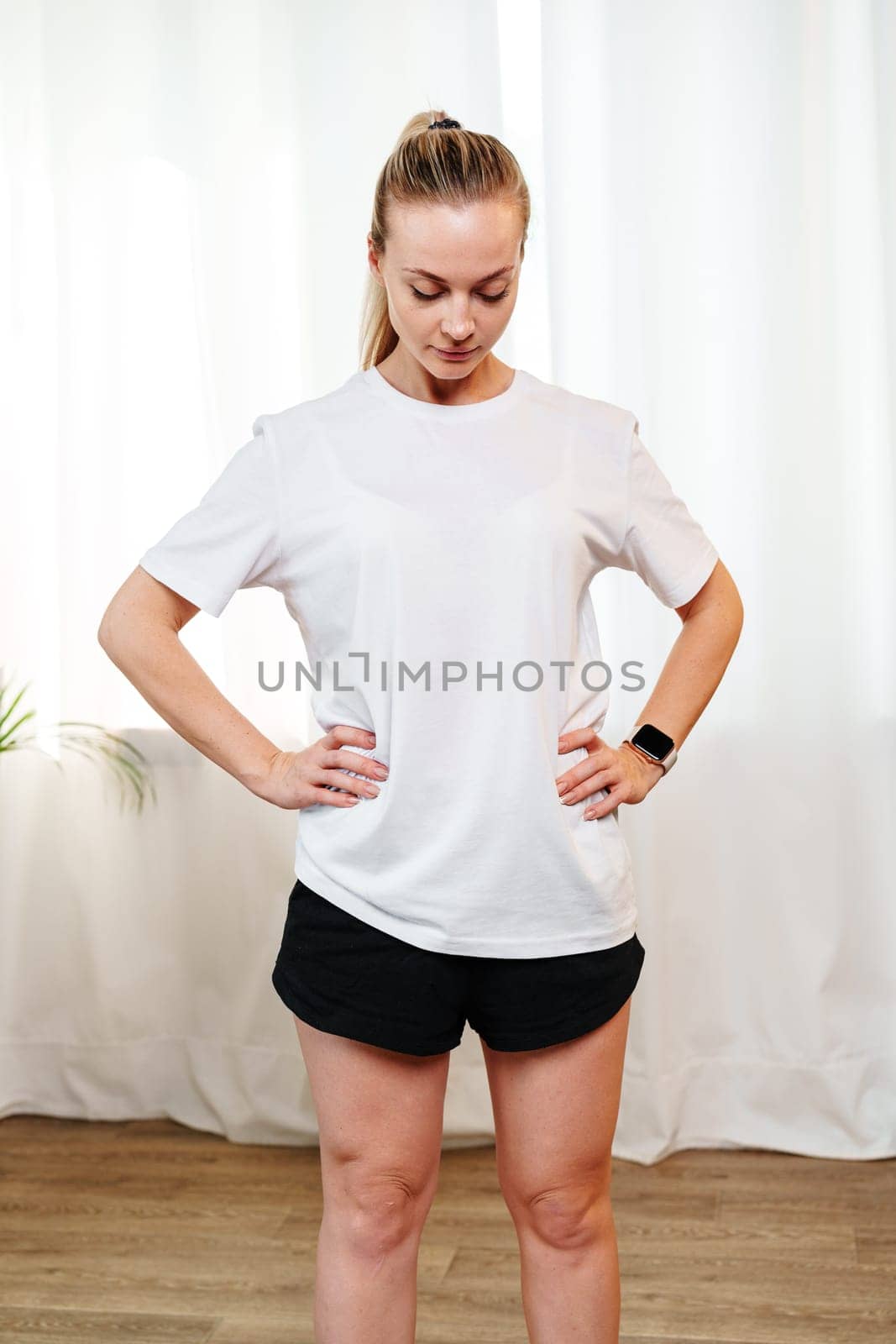 A woman stretches her neck and shoulders in a bright room surrounded by indoor plants during a home workout