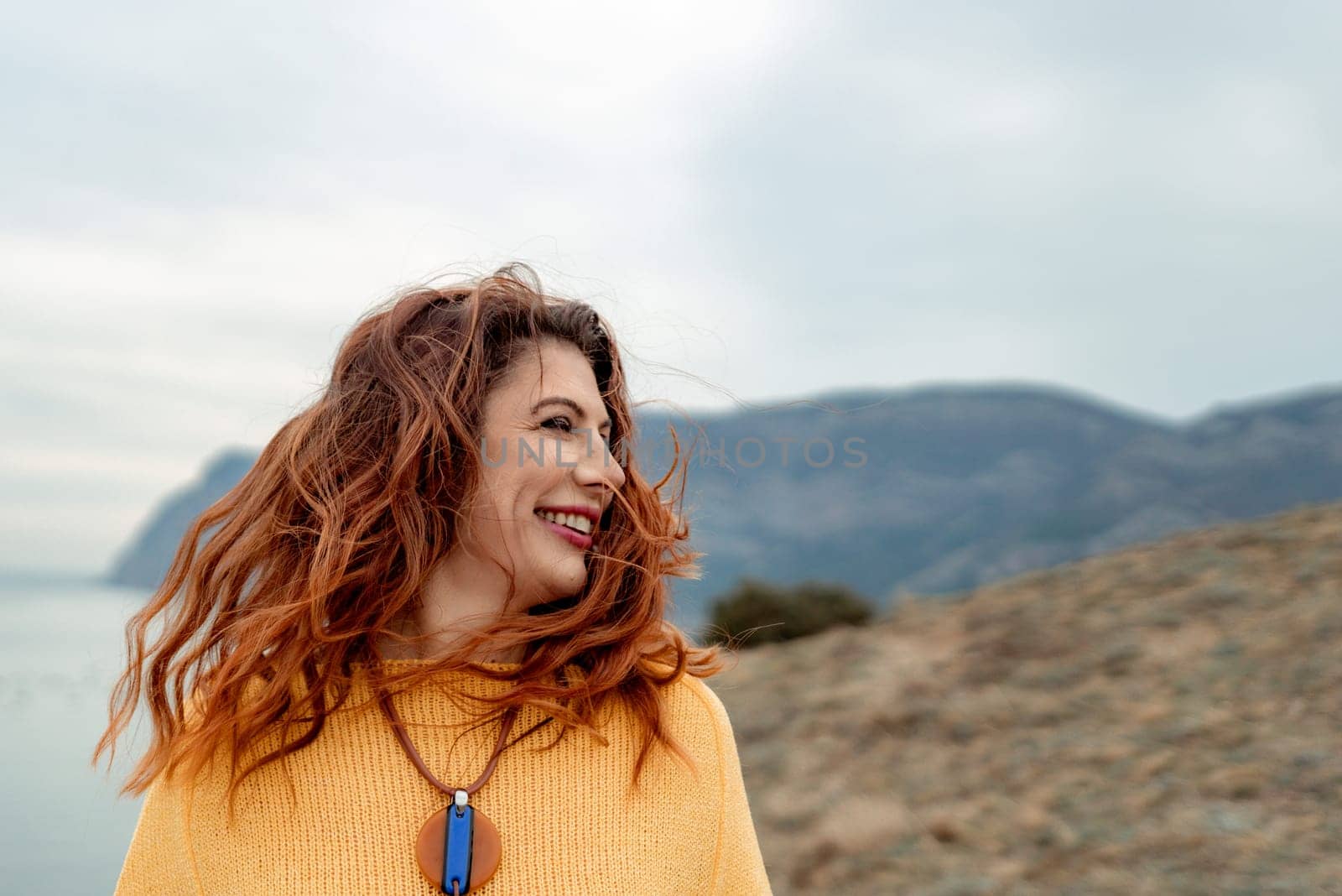 Portrait windswept hair happy woman against a backdrop of mountains and sea. Daylight illuminates the tranquil outdoor setting.