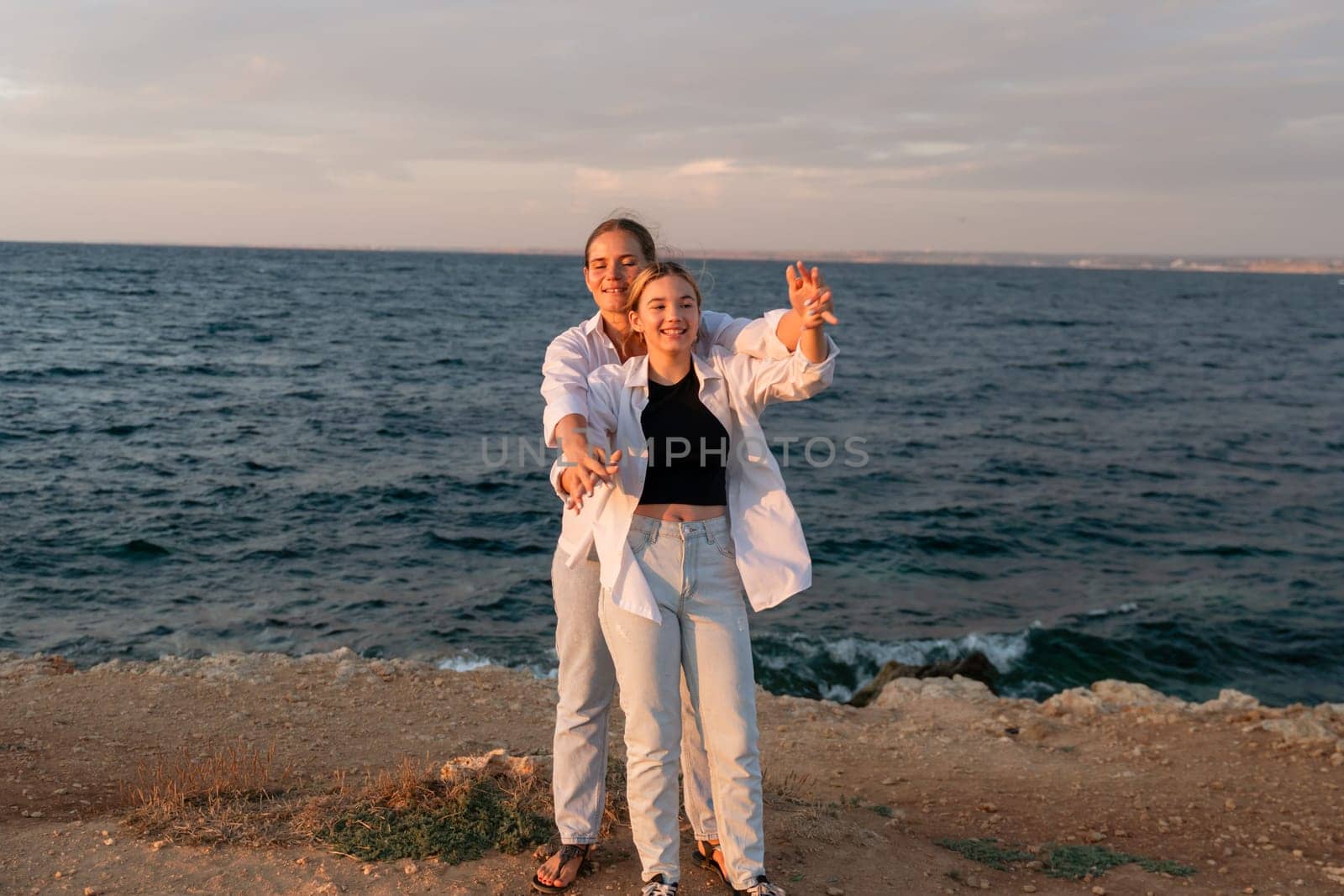 A couple takes a selfie on a rocky shoreline overlooking the ocean at sunset. The man is wearing a white shirt and the woman is wearing a black top and white pants.