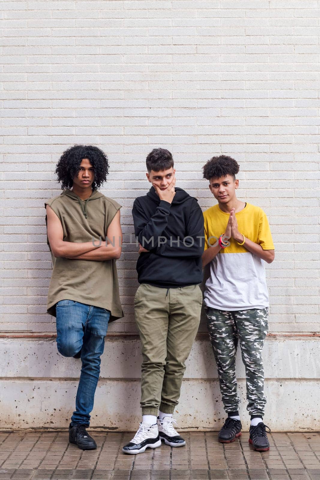 full body of multiethnic teenager friends with folded hands leaning against white brick wall, concept of friendship in youth and urban lifestyle, copyspace for text