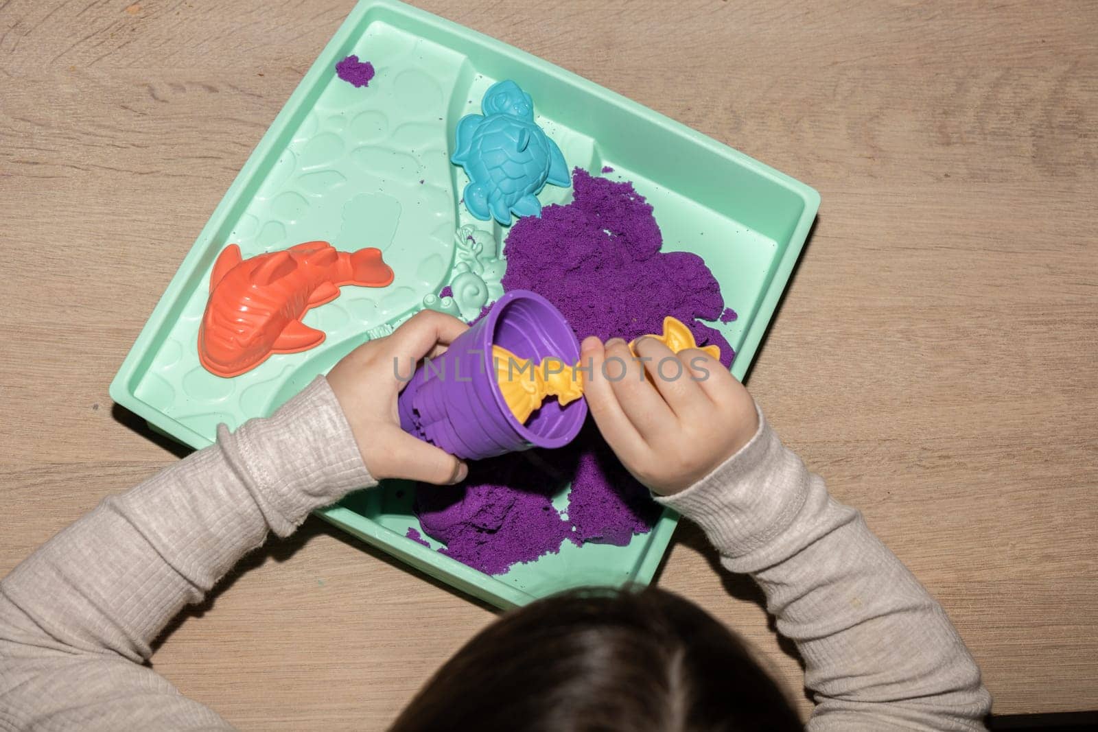 A child is using their fingers to create art with violet and magenta kinetic sand on a table. The leisure activity involves sharing gestures and thumb movements
