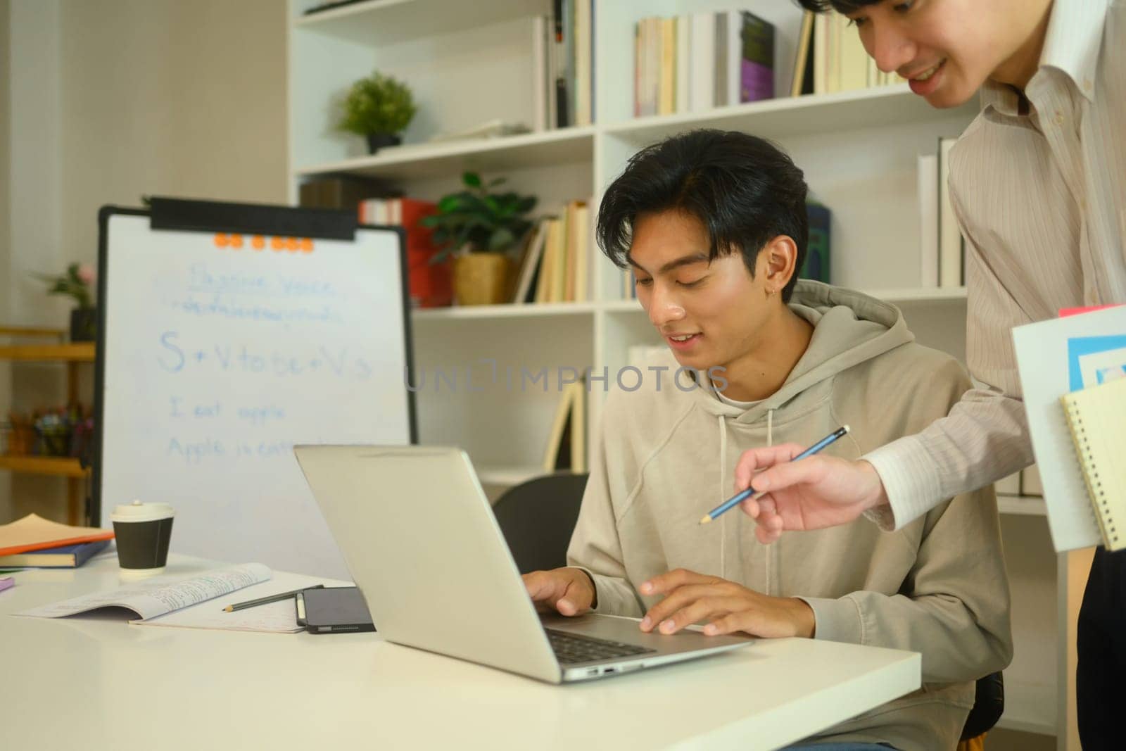 Asian male student in a hoodie working on a laptop, guided by a tutor at home.